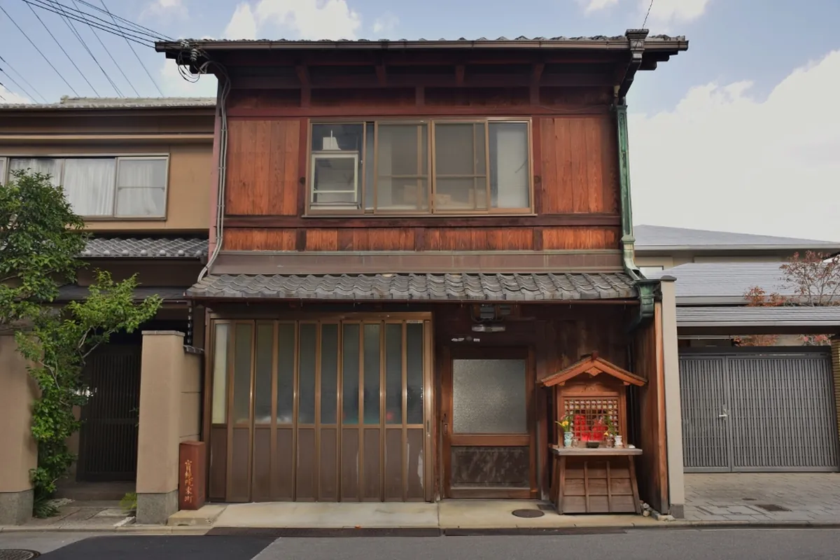 A two-storey wooden machiya townhouse on a Kyoto side street, with a tiled eave, timber cladding, a sliding glass door and a small roadside shrine beside it